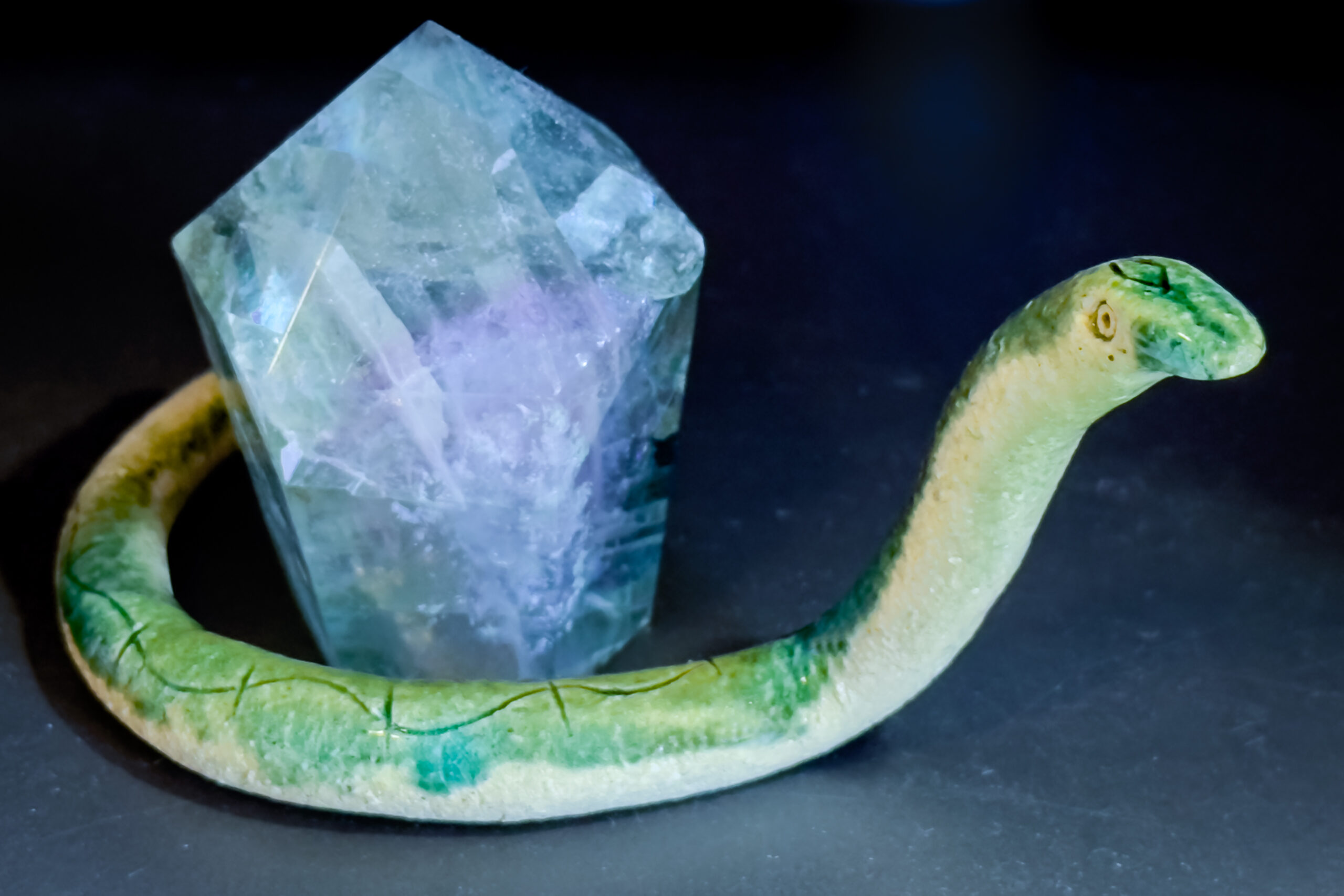 A green-glazed ceramic snake with its head raised. It is curned around a green and purple pentagon-shaped crystal