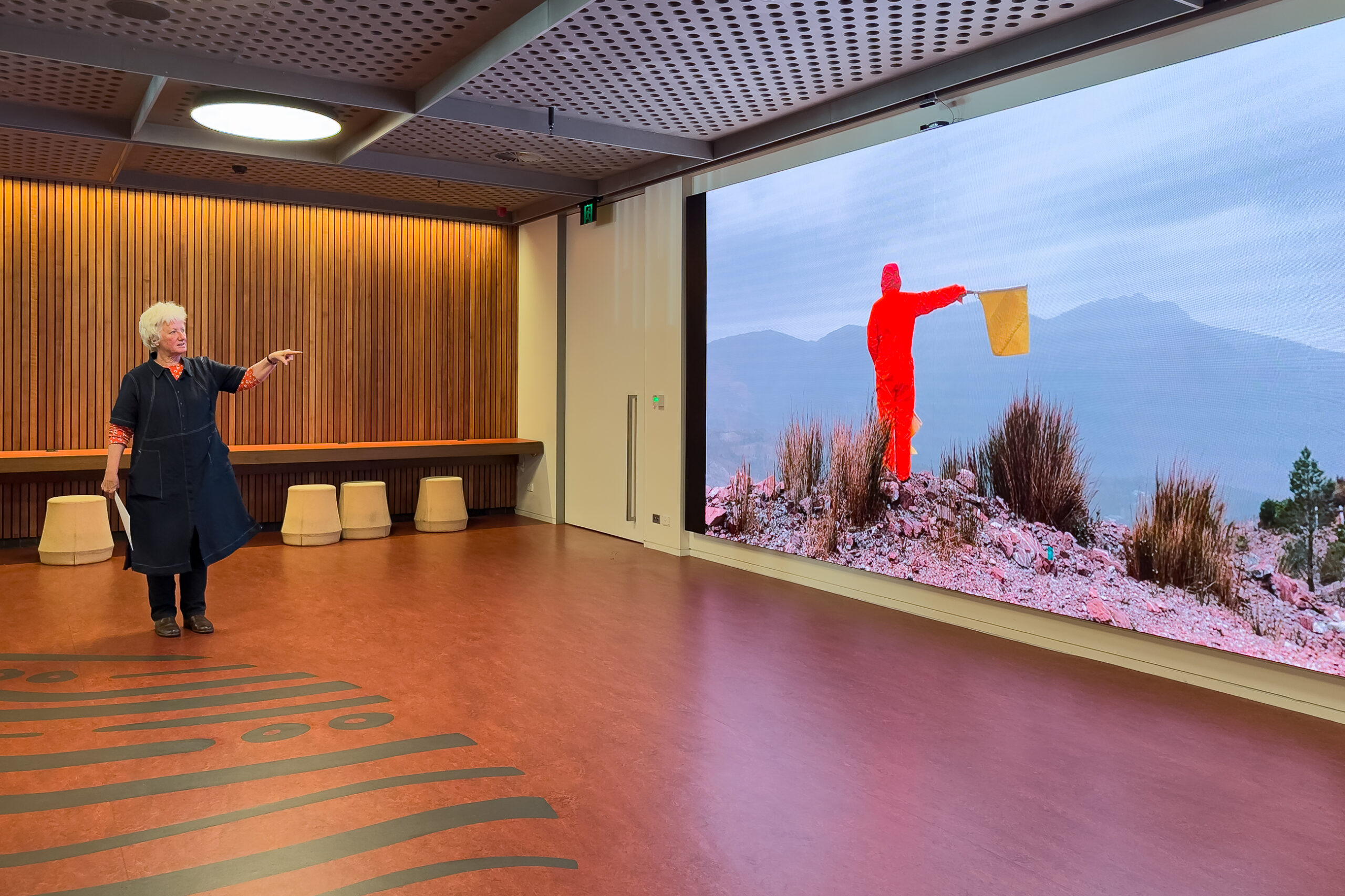 A woman is standing in front of a screen pointing to the image on the screen, which is a person dressed in bright orange holding out a small flag