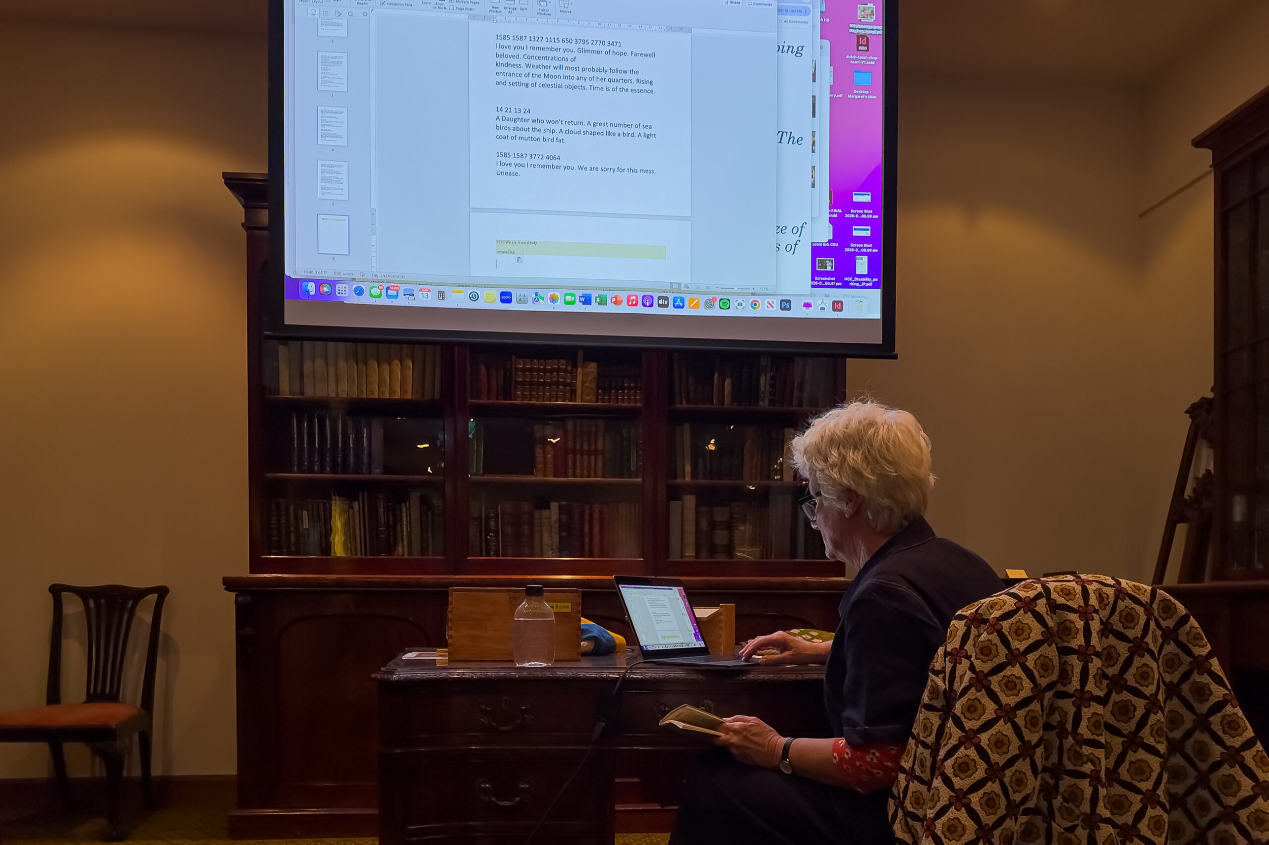 A woman sitting in front of a laptop on an old fashioned desk in room furnished with an old bookcase. Her work is also displayed on the large screen on the wall