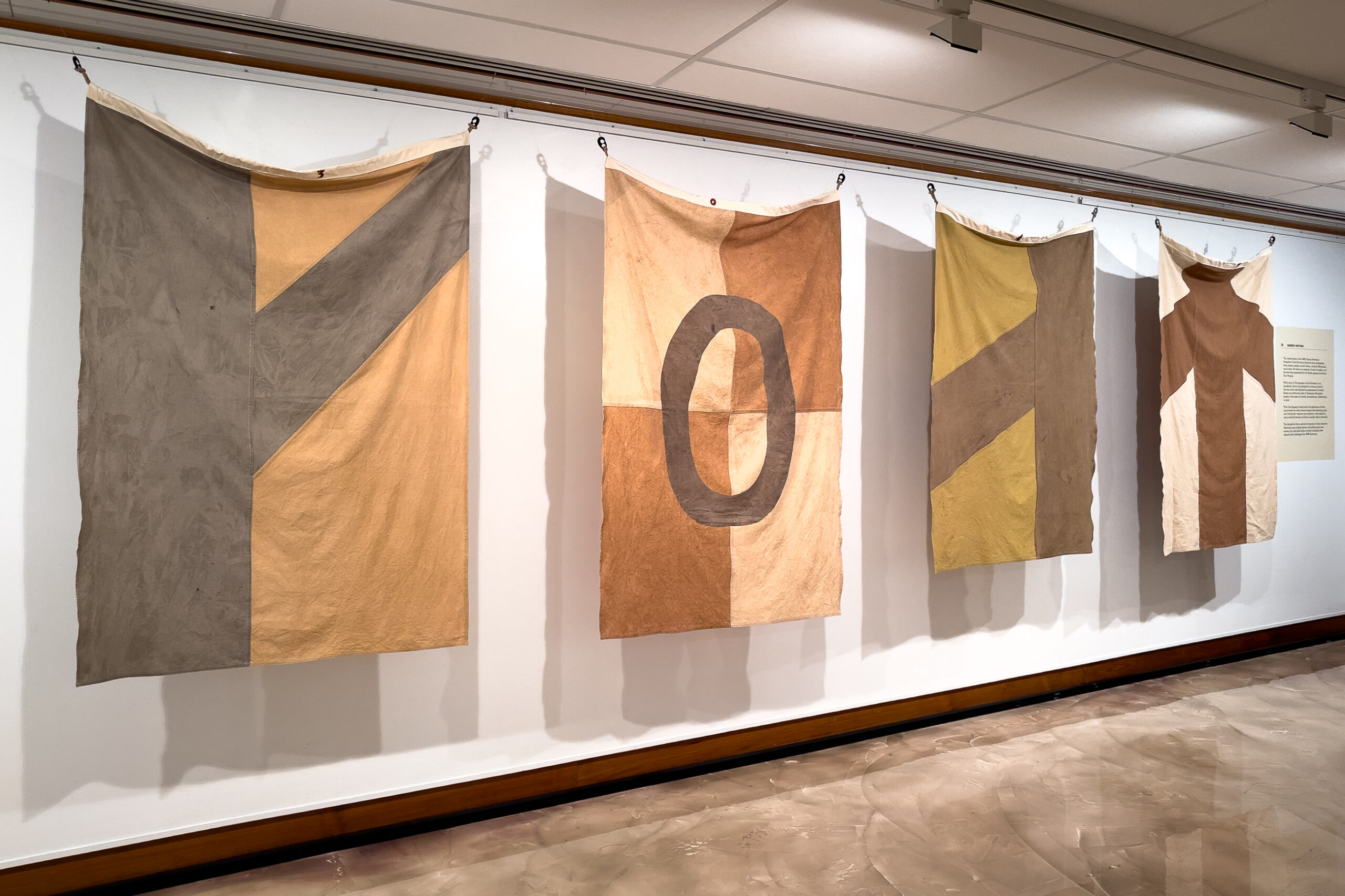 Four large flags in colours of brown and yellow hang on a gallery wall