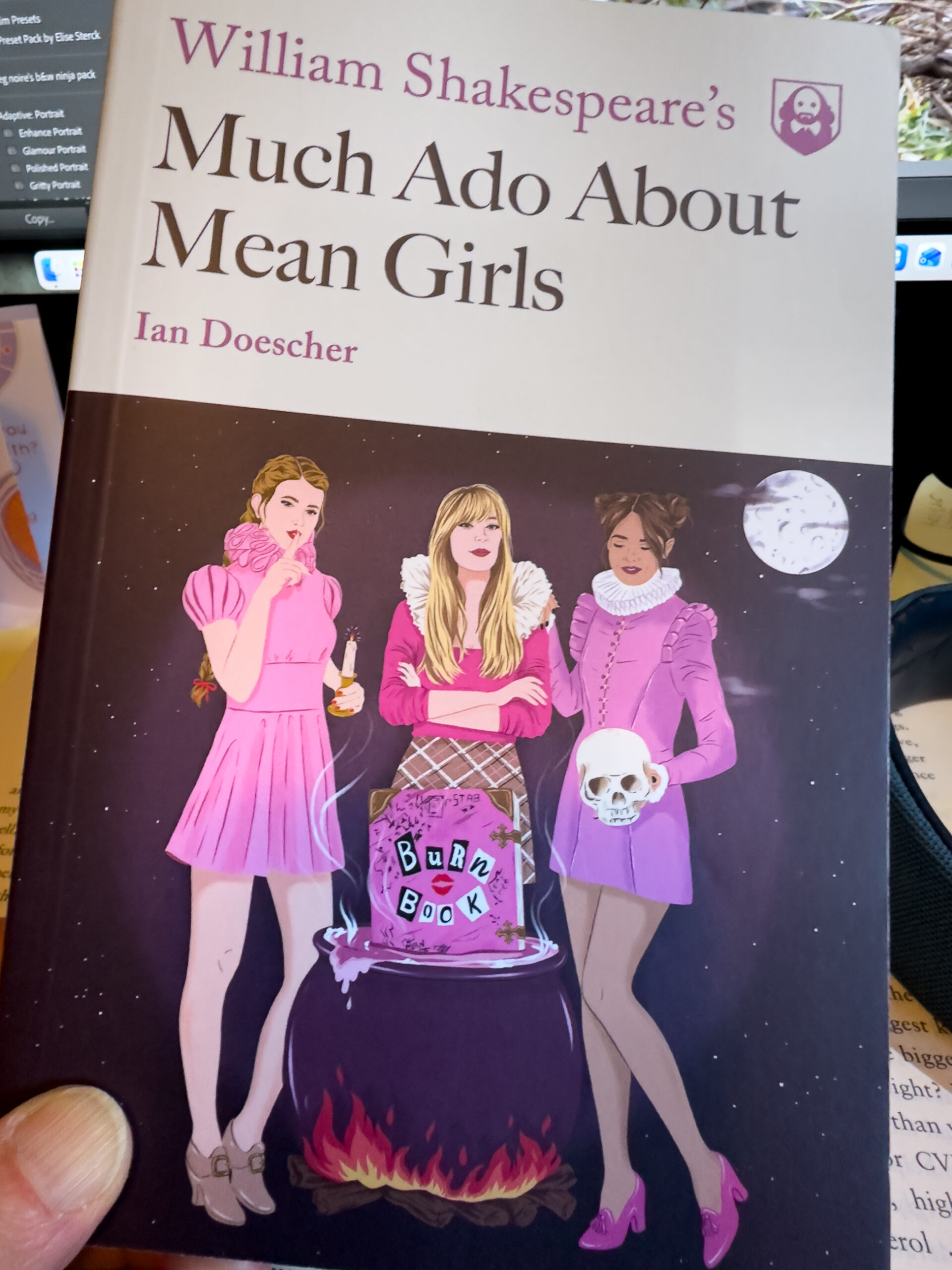 A book called “William Shakespeare’s Much Ado About Mean Girls”. The cover of the book features three girls dressed in pink next to a cauldron. One is holding a skull.