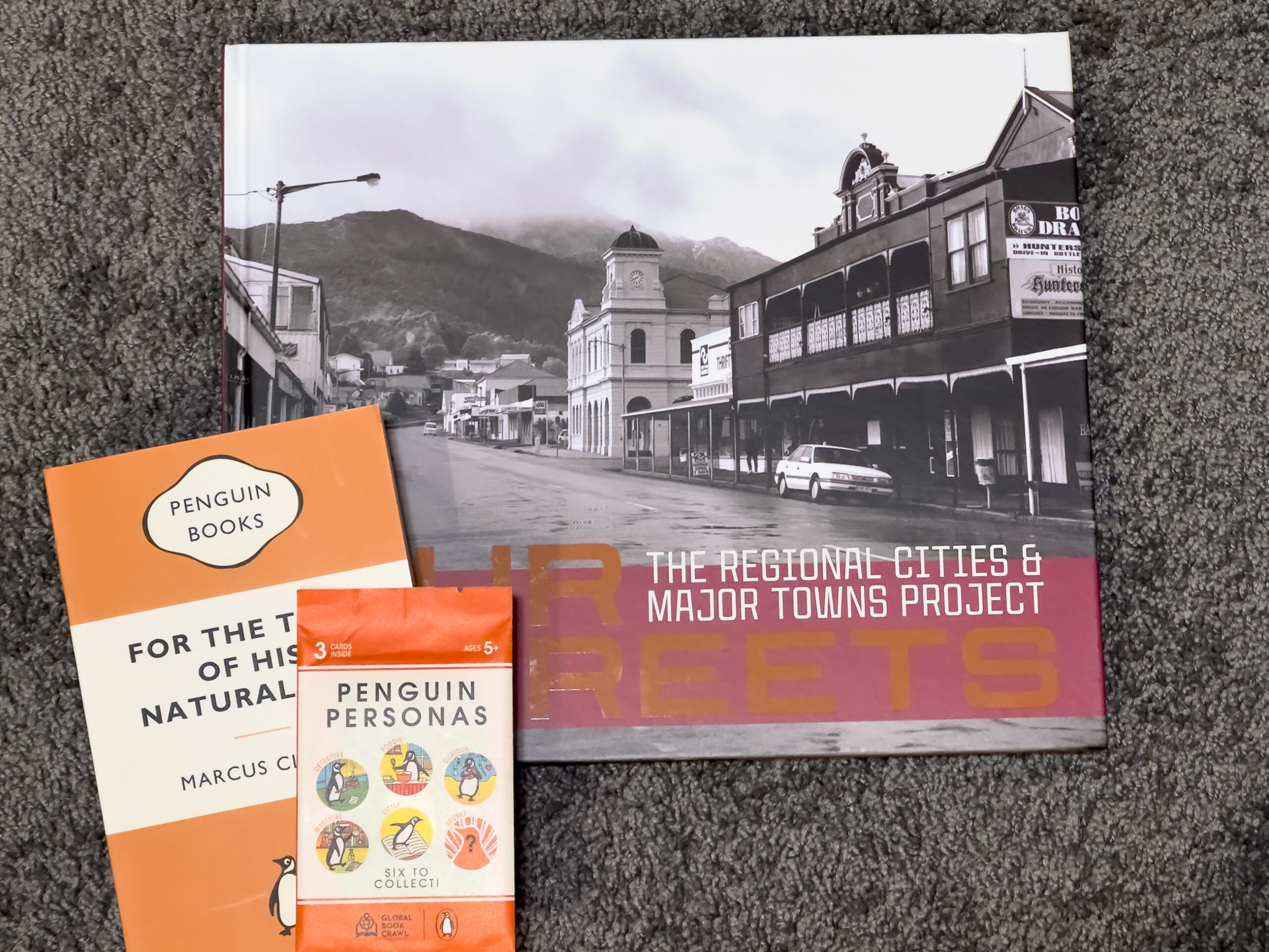 A large book with a black and white photo of a townscape called Our Streets: The Regional Cities and Major Towns Project and a smaller orange and hite book called For The Term of His Natural Life, and a little packet of "Penguin personas" cards on top