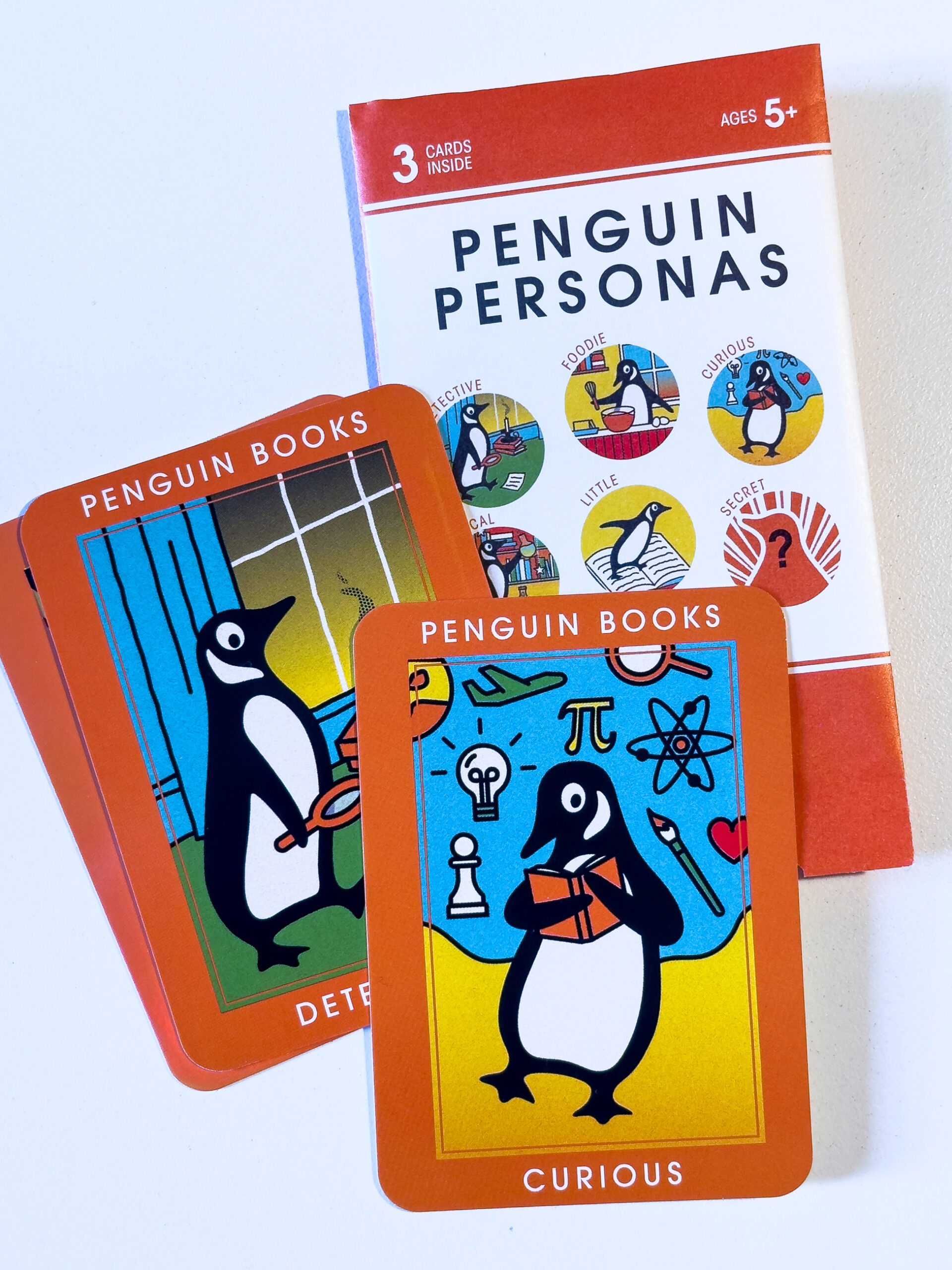 A set of thee cards sitting on top of a card packet. The top card is a picture of a curious penguin holding a book surrounded by many symbols. The second is the detective card, which has a penguin with a magnifying glass