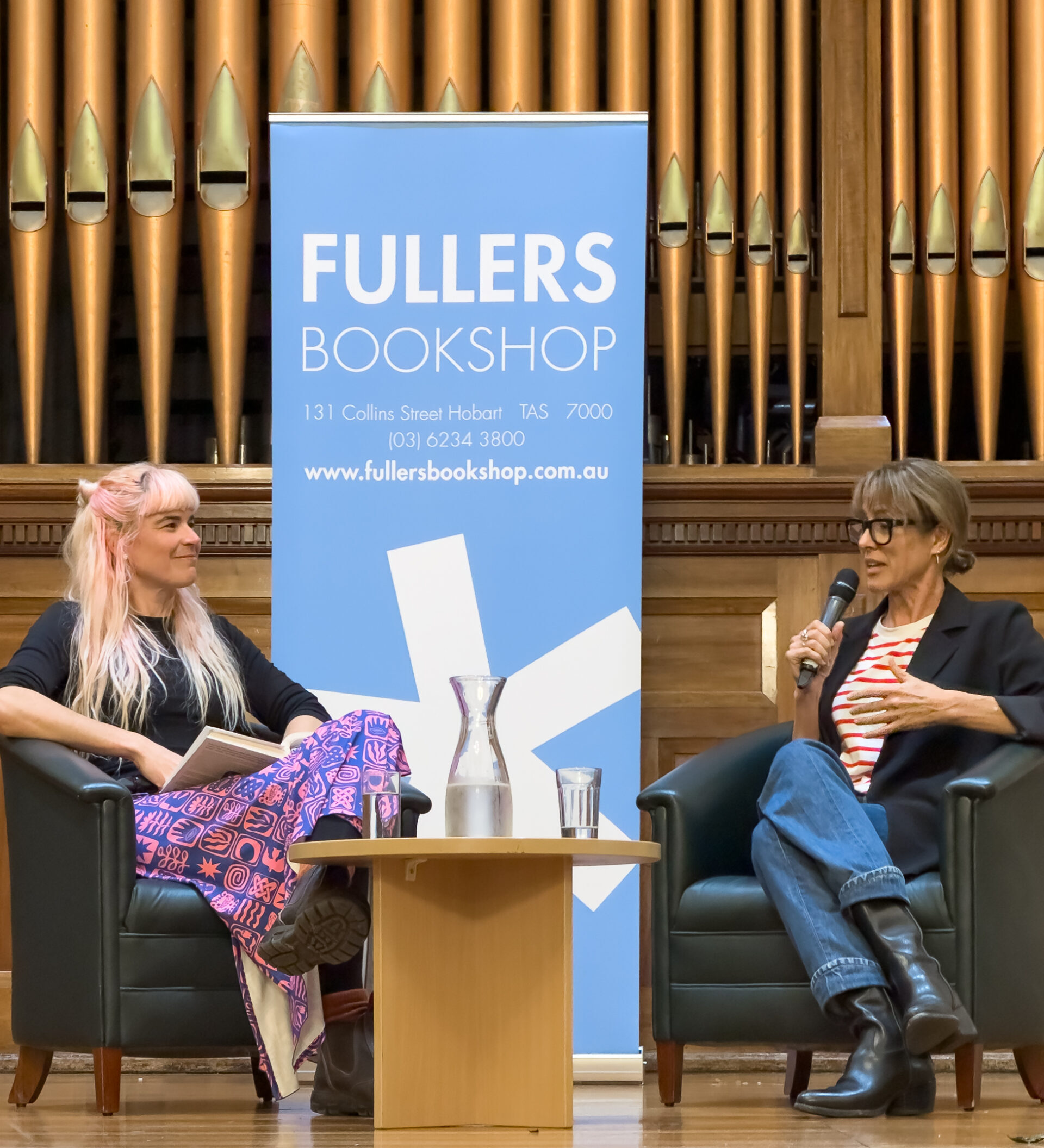 Two women sitting on chairs on a stage with a sign for Fullers Bookshop behind them