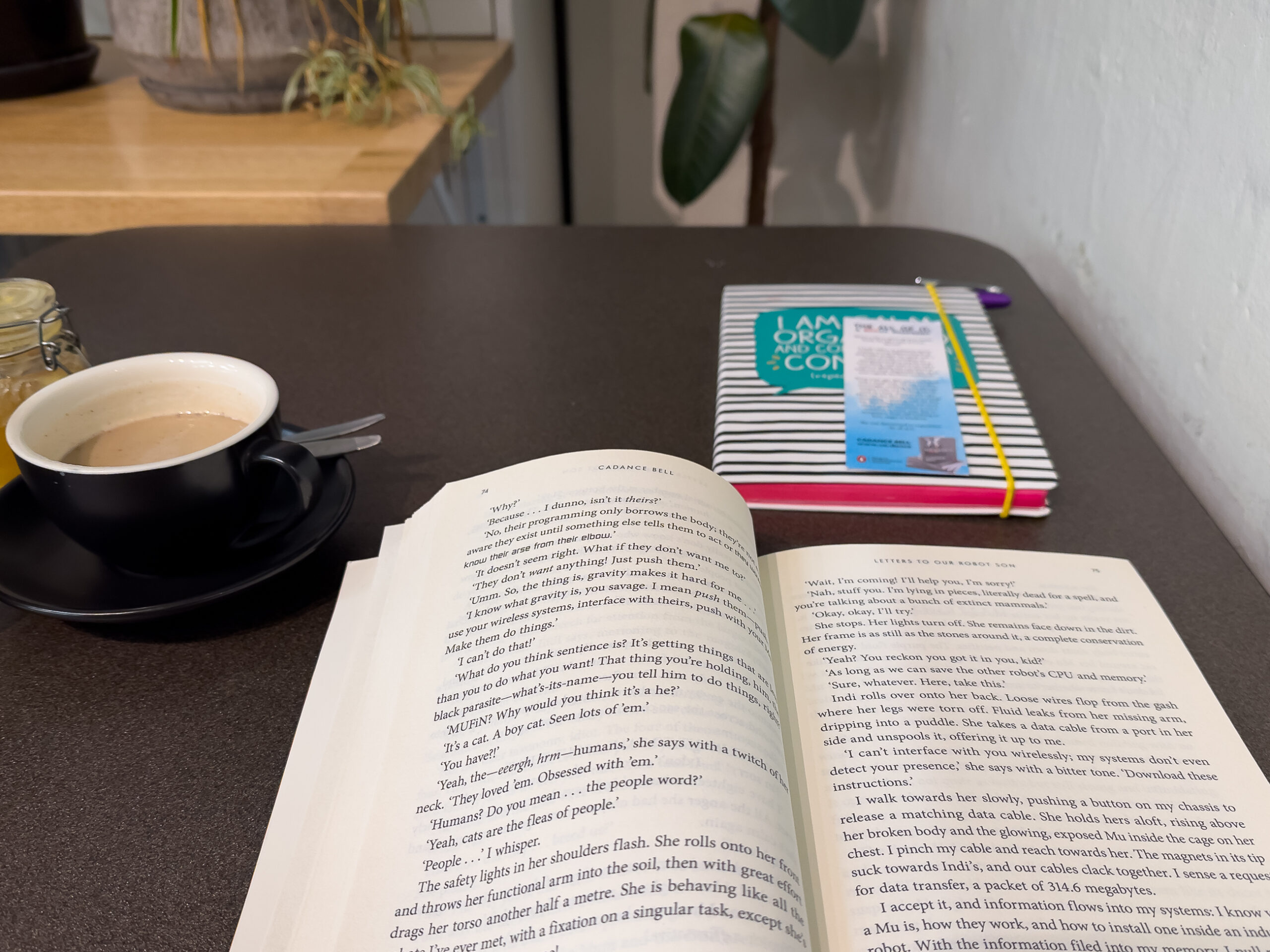 A book is open on a table. There is a cup of tea next to it on the left and a notebook also sitting on the table