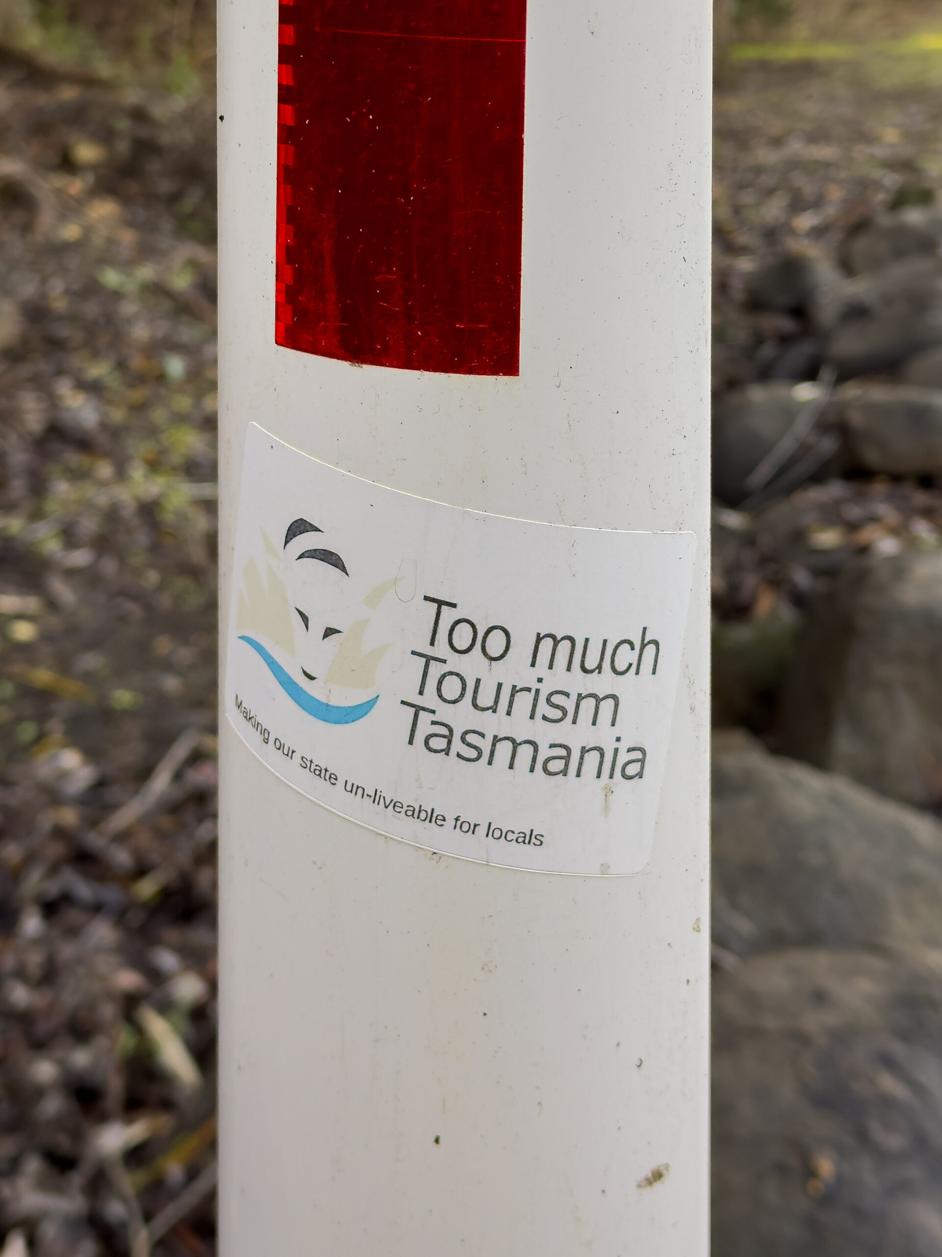 A sticker on a white post that says "too much Tourism Tasmania. Making our state unliveable for locals" There is a graphic of an old version of the state government logo
