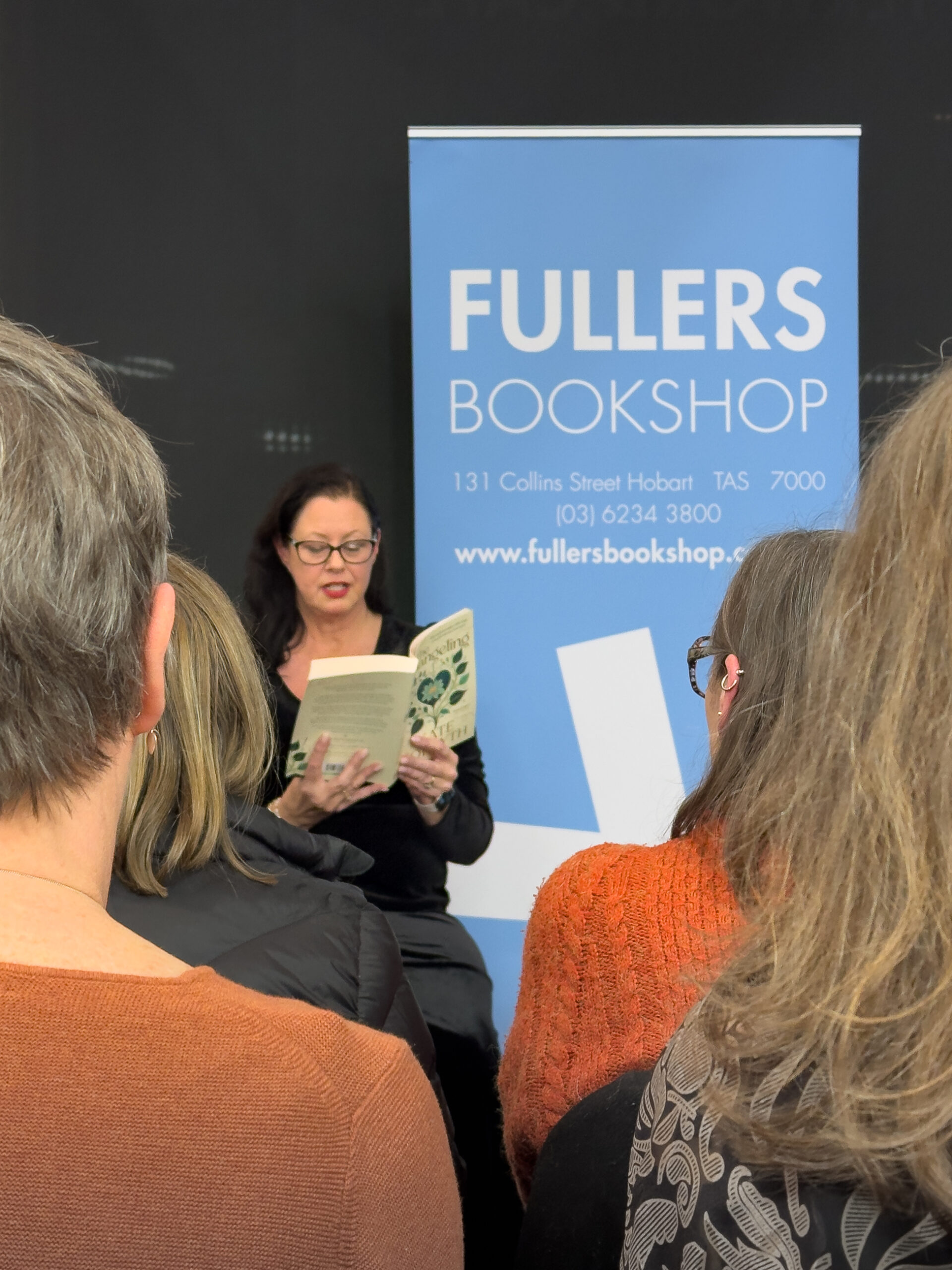 A woman with dark hair is reading from a book to a crowd of people. She is sitting near a blue sign with the words "Fullers Bookshop"