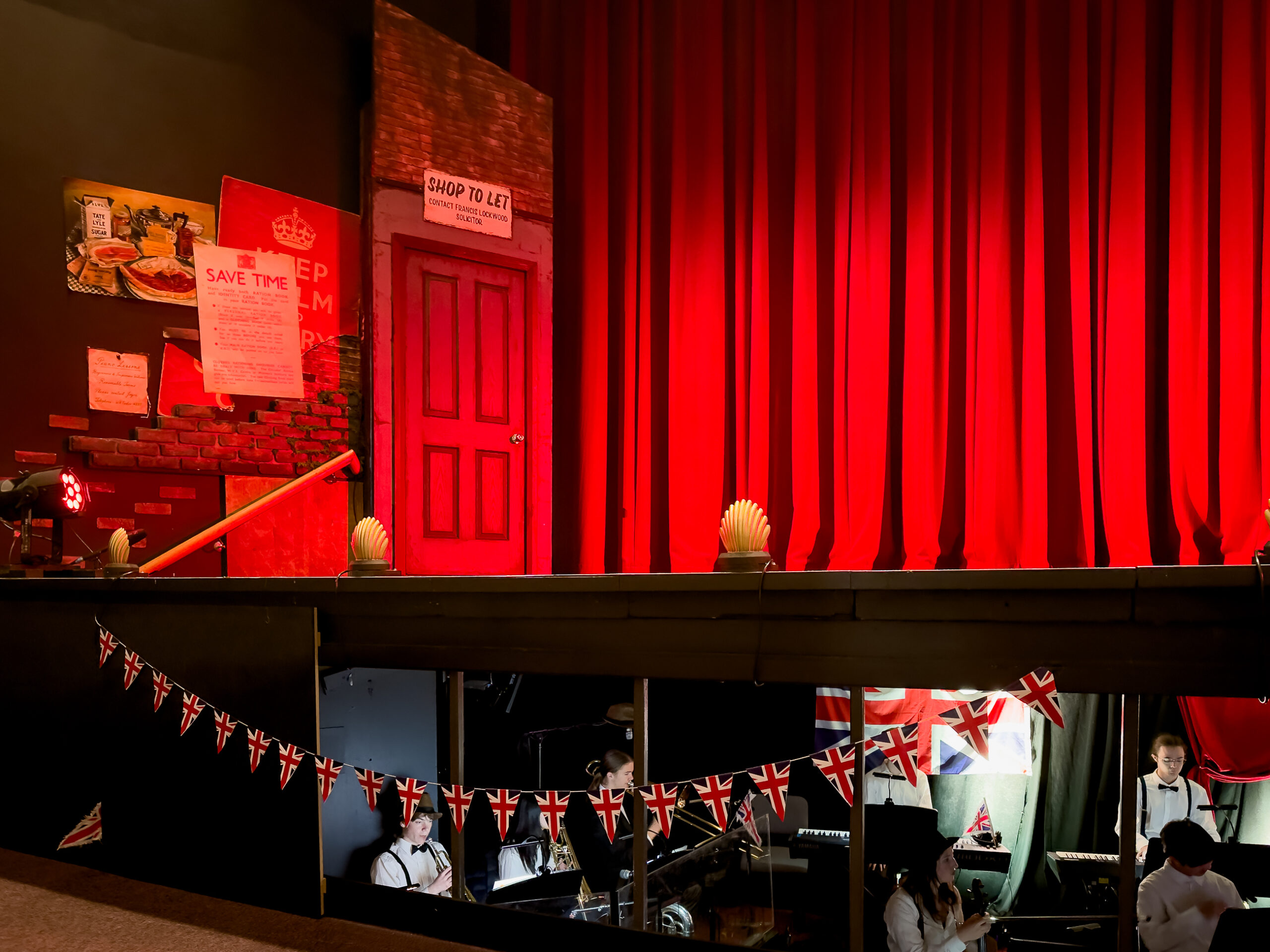 A stage set for the show Betty Blue Eyes. It's a red stage and there is bunting and doors of shops. The orchestra is in the pit underneath the stage
